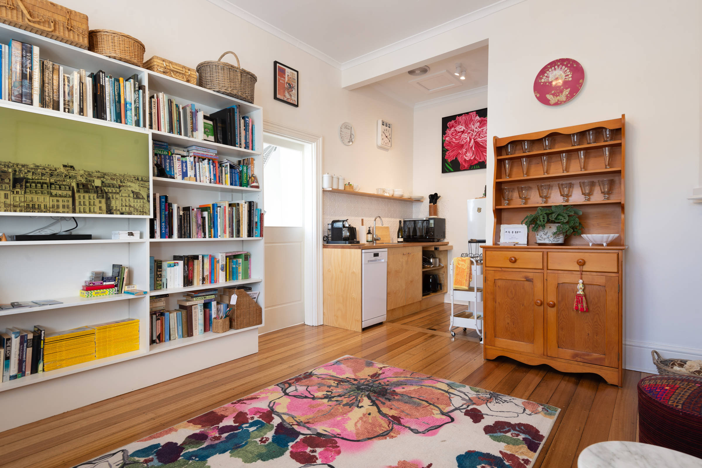 A small, tidy living space with a bookshelf, colourful floral rug, wooden cabinet, and compact kitchen area with appliances and art on the walls. Photo: Oliver Berlin.