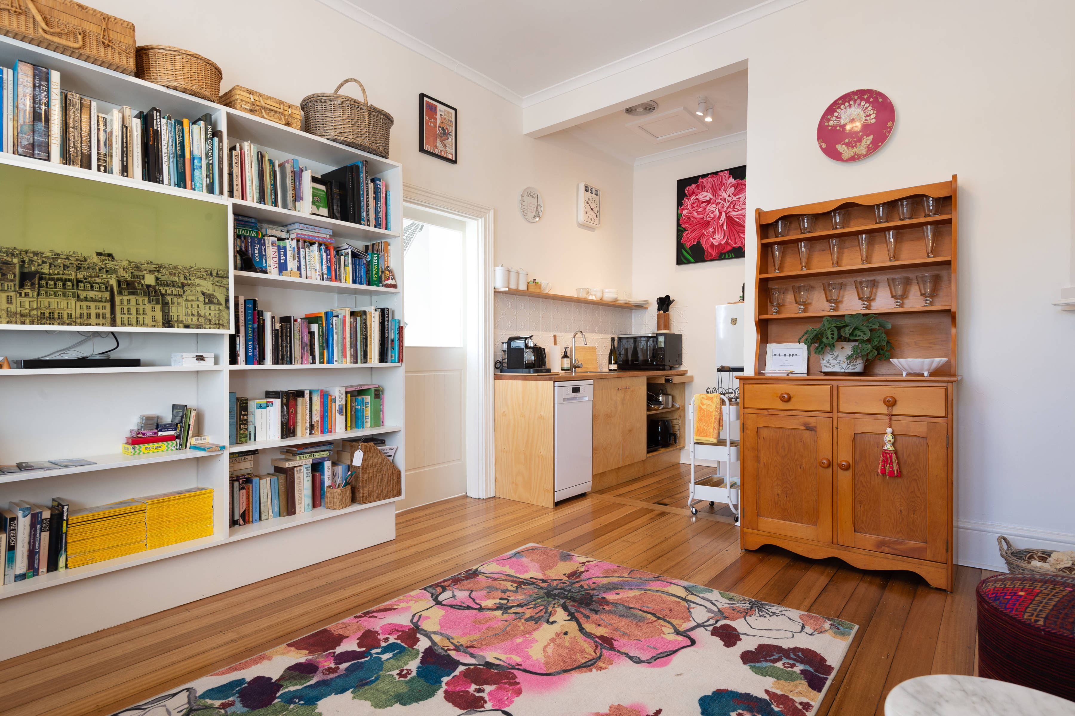 A small, tidy living space with a bookshelf, colourful floral rug, wooden cabinet, and compact kitchen area with appliances and art on the walls. Photo: Oliver Berlin.