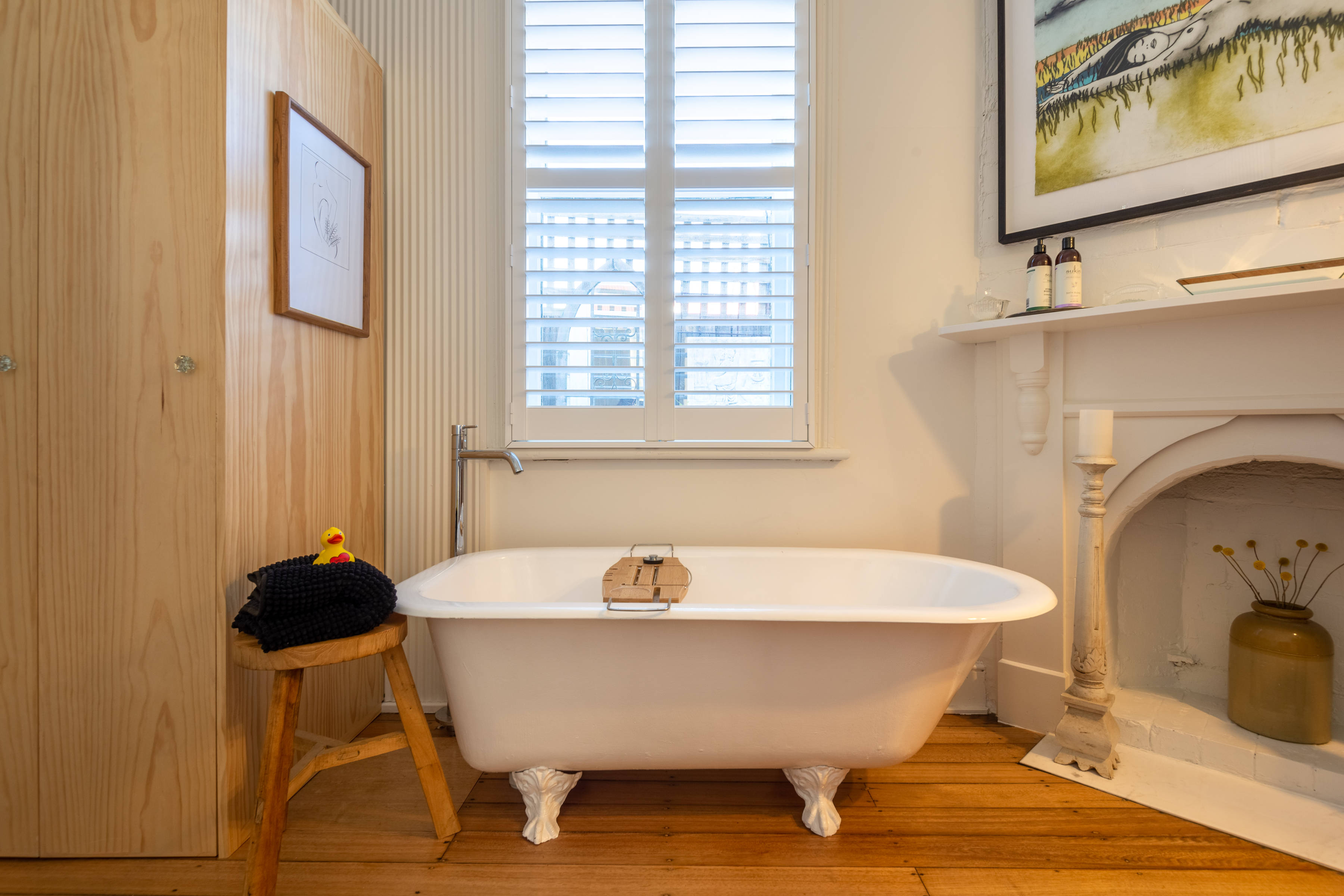 Claw foot bath top under window with stool and towel, fireplace, shelf, art work and laundry cupboard. Photo: Oliver Berlin.