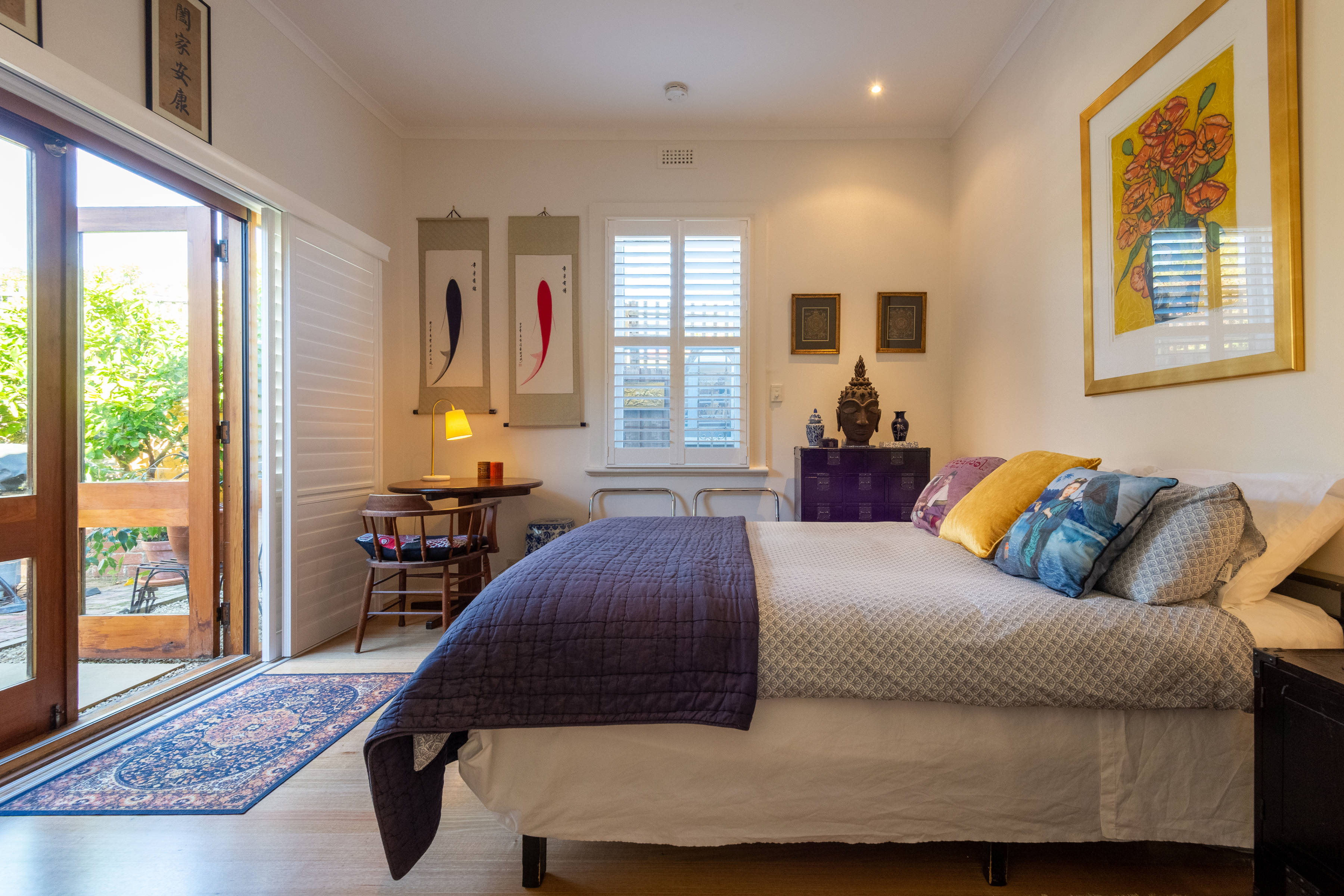 Queen bed with linen and cushions, desk, lamp, draws with ornaments, rug, door opening onto exterior. Photo: Oliver Berlin.